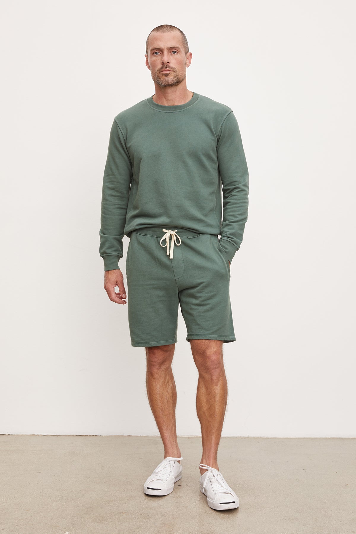 A man stands against a plain background, wearing a green sweatshirt, French terry Beckett shorts from Velvet by Graham & Spencer, and white sneakers.-36918621601985