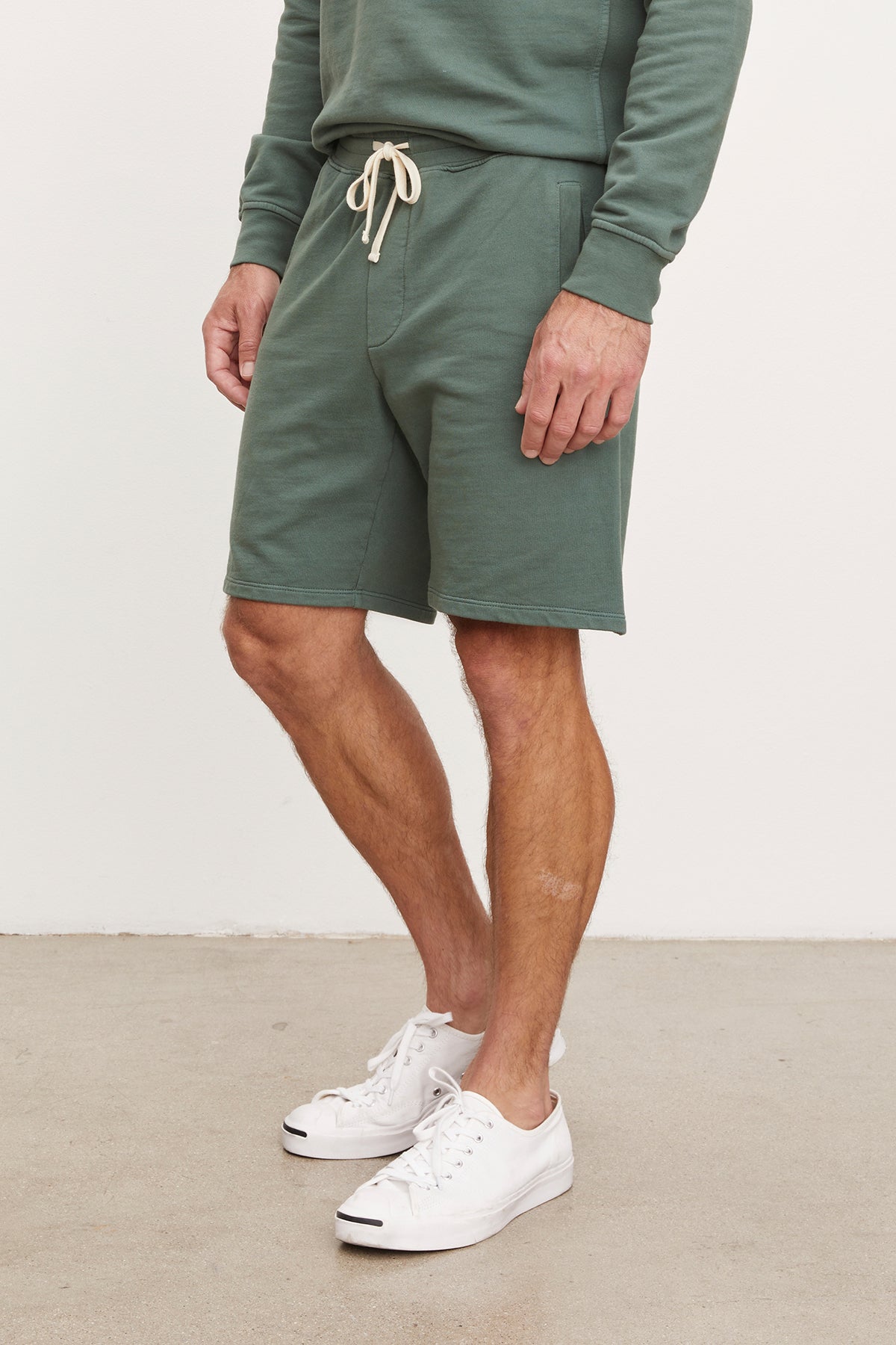 A man wearing a green sweatshirt and matching Velvet by Graham & Spencer Beckett shorts with white sneakers, cropped to show from the chest down.-36918621634753