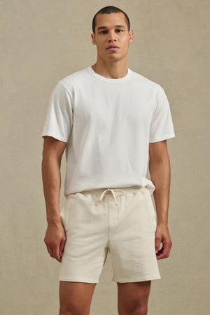 A person stands against a plain background, wearing a white short-sleeve t-shirt and the BOSLOW SHORT by Velvet by Graham & Spencer—relaxed fit shorts with a drawstring waist.