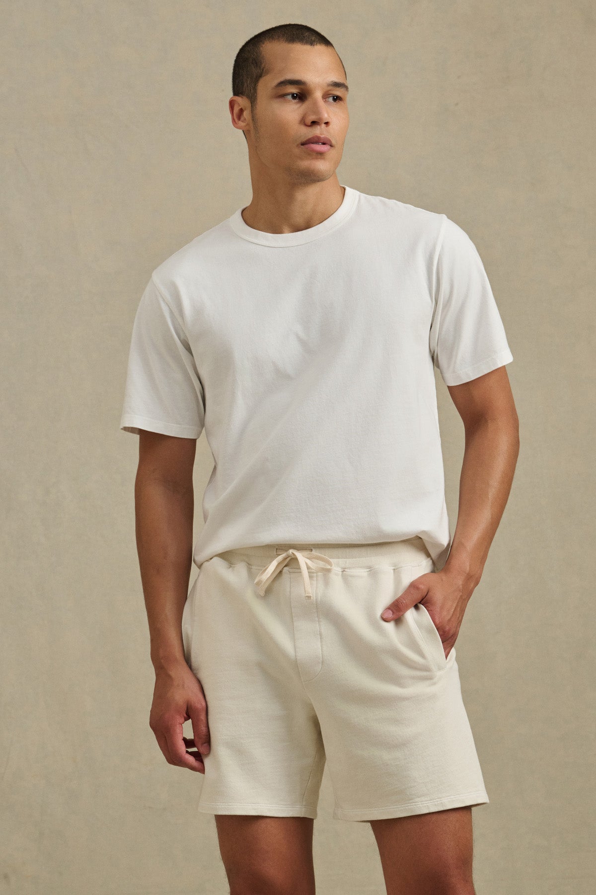 A man wearing a plain white t-shirt and Velvet by Graham & Spencer’s BOSLOW SHORT in relaxed fit, light beige French terry stands against a neutral background, perfect for everyday wear.-41374357094593