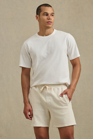 A man wearing a plain white t-shirt and Velvet by Graham & Spencer’s BOSLOW SHORT in relaxed fit, light beige French terry stands against a neutral background, perfect for everyday wear.