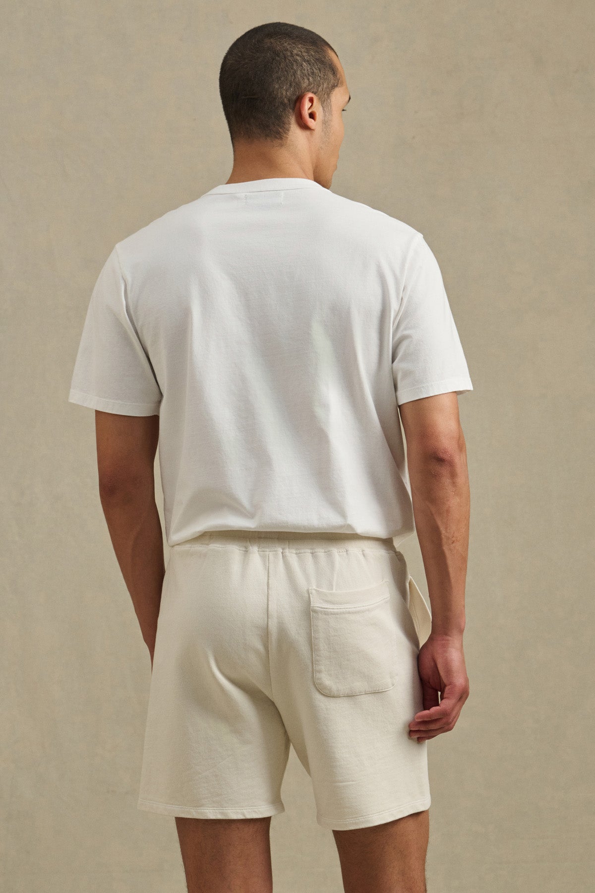 A man faces away, wearing Velvet by Graham & Spencer's BOSLOW SHORT—cream-colored French terry shorts with a back pocket, perfect for everyday wear—paired with a plain white tee against a neutral background.-41374357225665