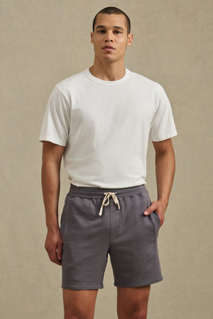 A man wearing a plain white t-shirt and Velvet by Graham & Spencer BOSLOW SHORT—relaxed fit French terry shorts with a drawstring waist—stands with one hand in his pocket against a neutral background.