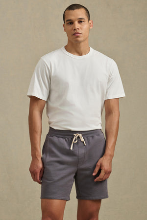 A man stands against a plain background wearing a white short-sleeve t-shirt and gray BOSLOW SHORTS by Velvet by Graham & Spencer, featuring a drawstring waist.