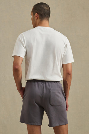 A person faces away, wearing Velvet by Graham & Spencer's BOSLOW SHORT—relaxed fit French terry shorts with drawstring waist and back pocket—paired with a plain white t-shirt, set against a neutral background.