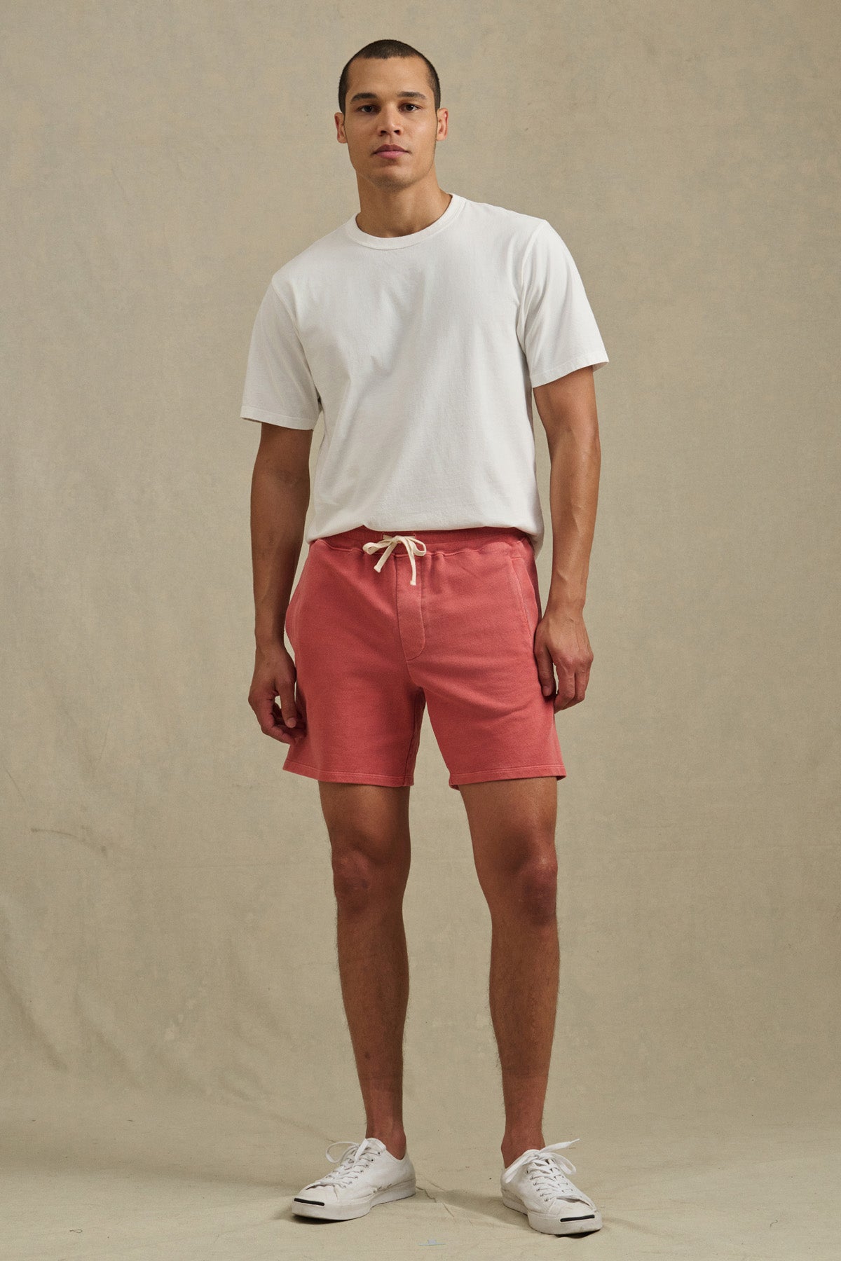 A man stands against a plain background wearing the BOSLOW SHORT in pink by Velvet by Graham & Spencer, styled with a white t-shirt and white sneakers.-41428357316801