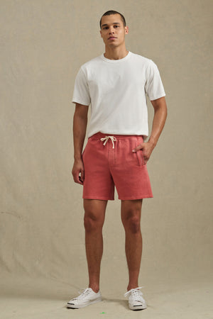 A man stands against a plain backdrop wearing a white t-shirt, white sneakers, and Velvet by Graham & Spencer BOSLOW SHORT—relaxed fit pink French terry shorts with a drawstring elastic waist.