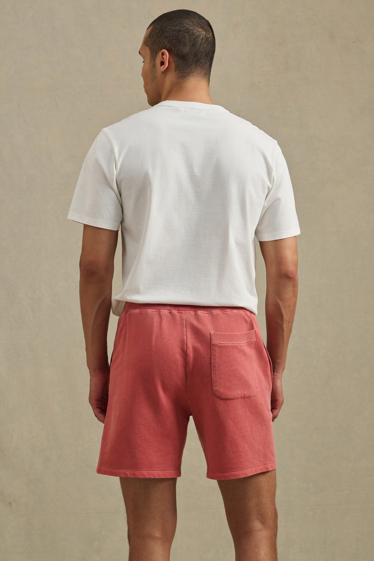 A man faces away, wearing a white T-shirt and Velvet by Graham & Spencer BOSLOW SHORT—relaxed fit pink French terry shorts with a back pocket—against a plain beige background.