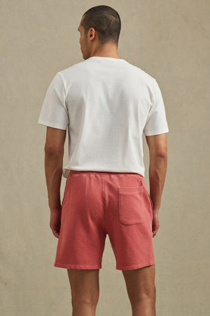 A man faces away, wearing a white T-shirt and Velvet by Graham & Spencer BOSLOW SHORT—relaxed fit pink French terry shorts with a back pocket—against a plain beige background.