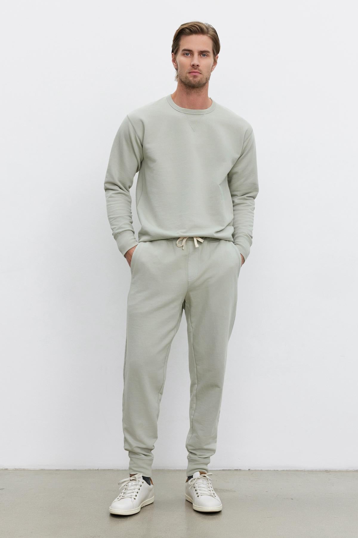   A man stands against a plain white background wearing a light gray, long-sleeved sweatshirt made from mid-weight French terry and matching DUSTY SWEATPANT by Velvet by Graham & Spencer with an elastic drawstring waist and ribbed cuffs, paired with white sneakers. His hands are in his pockets. 