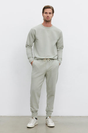 A man stands against a plain white background wearing a light gray, long-sleeved sweatshirt made from mid-weight French terry and matching DUSTY SWEATPANT by Velvet by Graham & Spencer with an elastic drawstring waist and ribbed cuffs, paired with white sneakers. His hands are in his pockets.