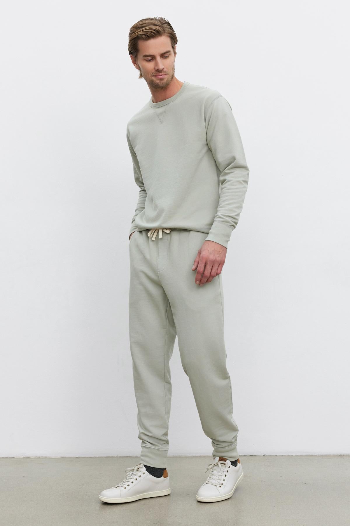   A man is standing against a plain white background, wearing a light gray sweatshirt, Velvet by Graham & Spencer DUSTY SWEATPANT with an elastic drawstring waist, and white sneakers. He has his hands in his pockets and is looking slightly downwards. 