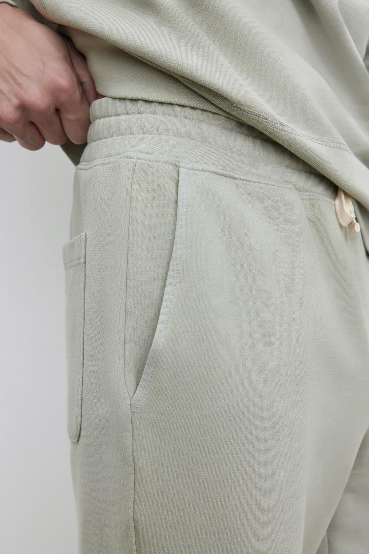   Close-up of someone wearing Velvet by Graham & Spencer DUSTY SWEATPANT made of soft fabric, featuring an elastic drawstring waist with one hand resting on the waistband. 