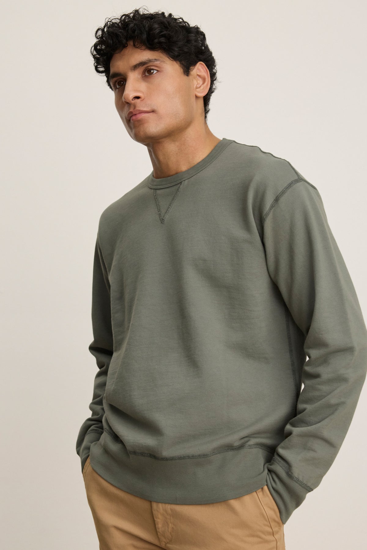 A man stands with his hands in his pockets against a neutral background, wearing the RAE SWEATSHIRT in olive green by Velvet by Graham & Spencer and tan pants.-39446044606657