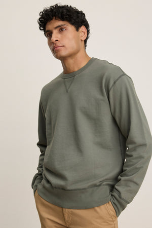 A man stands with his hands in his pockets against a neutral background, wearing the RAE SWEATSHIRT in olive green by Velvet by Graham & Spencer and tan pants.