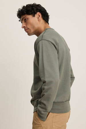 A man with short dark hair models the Velvet by Graham & Spencer RAE SWEATSHIRT in sage green loopback French terry, paired with beige pants, standing sideways against a plain light background.