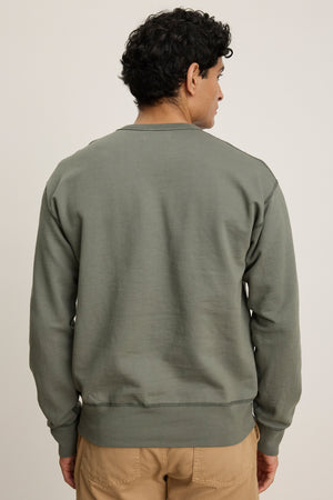 A man with short curly hair stands with his back to the camera, wearing the olive green RAE SWEATSHIRT by Velvet by Graham & Spencer, crafted from premium loopback French terry, and tan pants against a plain background.