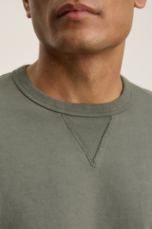 A close-up shows a person wearing the RAE SWEATSHIRT by Velvet by Graham & Spencer in olive green, featuring a classic crew neckline, with only the lower face and upper chest visible.