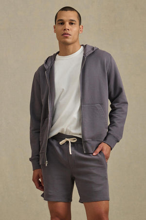 A person stands against a plain beige background wearing the VINCENT HOODIE by Velvet by Graham & Spencer, a vintage-inspired gray full-zip hoodie with matching drawstring shorts layered over a white t-shirt.