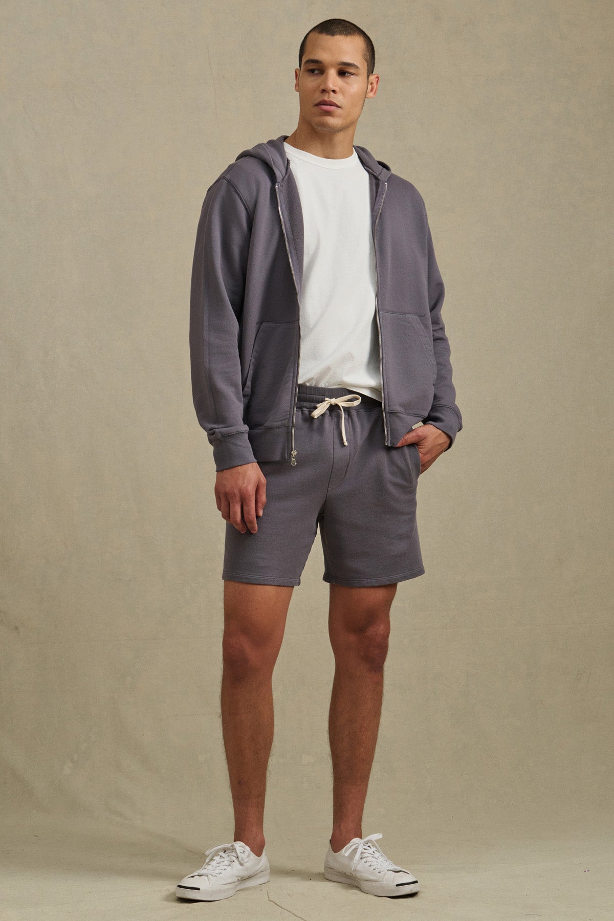 A man stands against a neutral background wearing Velvet by Graham & Spencer's BOSLOW SHORT, a relaxed fit French terry short with a drawstring waist, paired with a gray zip-up hoodie, white t-shirt, and white sneakers.-41626497450177