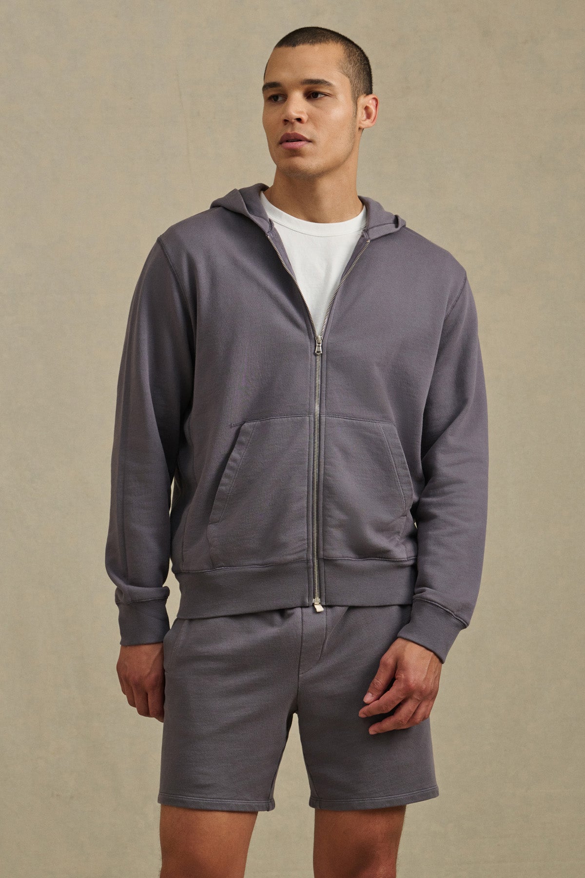A man wears the VINCENT HOODIE by Velvet by Graham & Spencer—a vintage-inspired light purple full-zip hoodie in soft French terry cotton—paired with a white t-shirt and matching shorts against a neutral background.-41626497482945