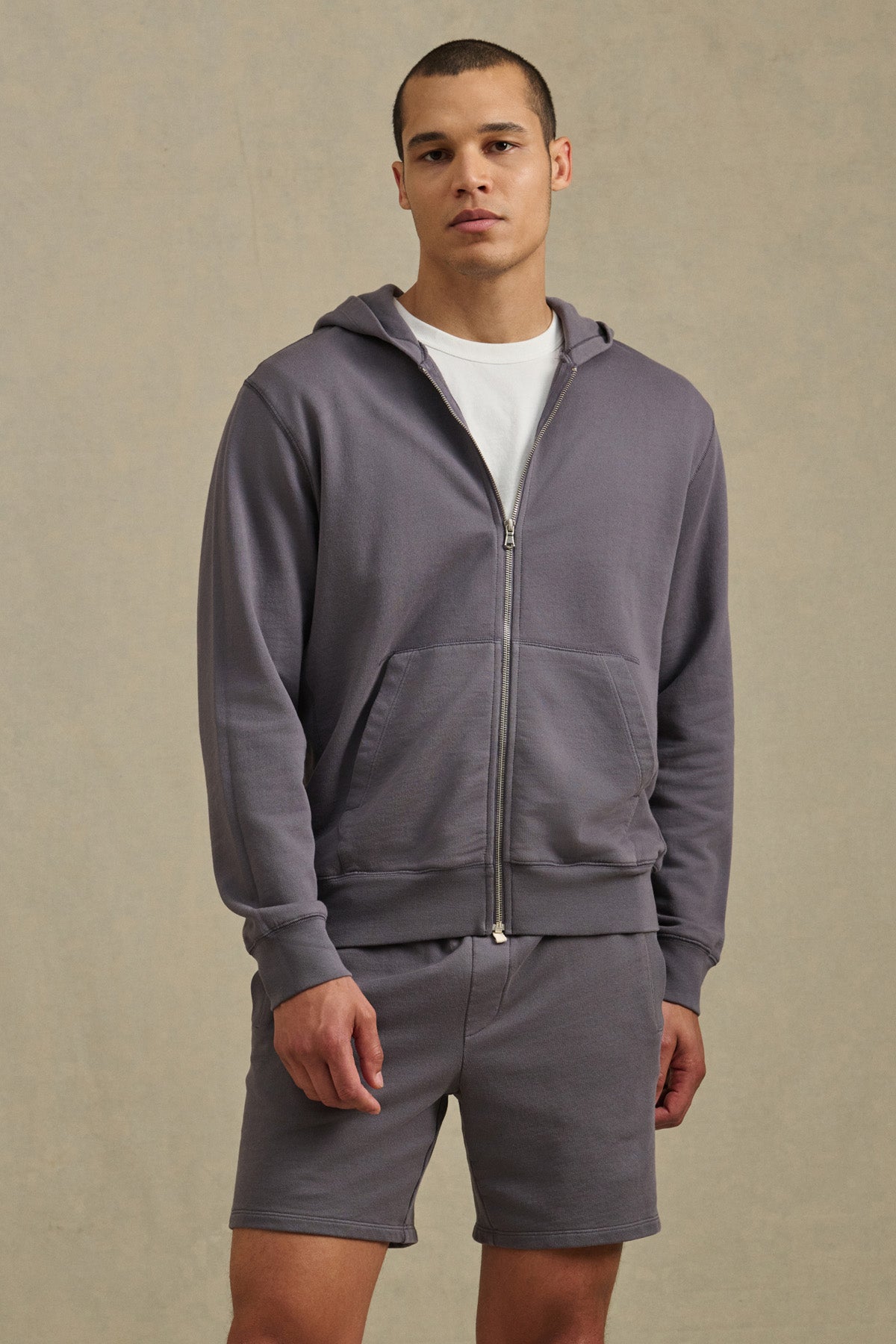 A man wears the VINCENT HOODIE by Velvet by Graham & Spencer— a vintage-inspired gray French terry cotton full-zip hoodie— with matching shorts and a white tee, standing against a neutral background, looking at the camera.