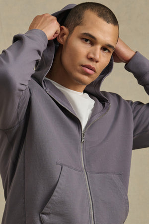 Wearing the VINCENT HOODIE by Velvet by Graham & Spencer, a person in gray French terry cotton with a white tee pulls up the hood and faces the camera against a neutral background.