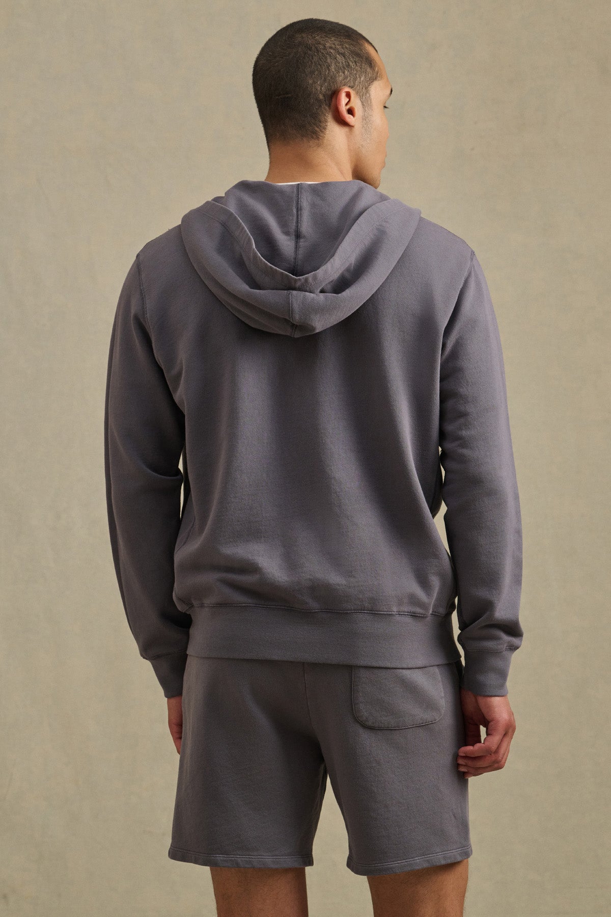 A man stands facing away, wearing the VINCENT HOODIE by Velvet by Graham & Spencer in vintage-inspired French terry cotton gray, along with matching gray shorts, set against a simple light background.-41626497581249