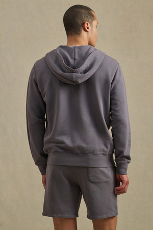 A man stands facing away, wearing the VINCENT HOODIE by Velvet by Graham & Spencer in vintage-inspired French terry cotton gray, along with matching gray shorts, set against a simple light background.