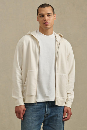 A person faces forward wearing the VINCENT HOODIE by Velvet by Graham & Spencer— a cream full-zip French terry cotton hoodie — layered over a white tee and blue jeans, set against a neutral background.