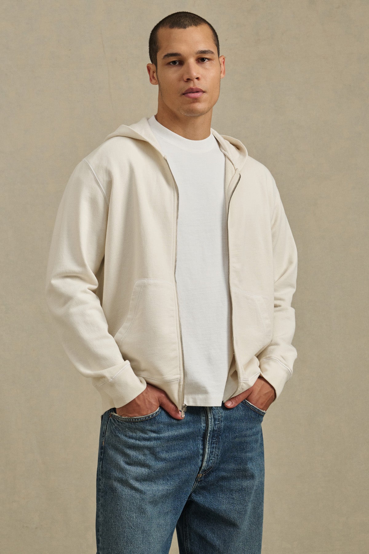 A person stands facing forward, hands in pockets, wearing the VINCENT HOODIE by Velvet by Graham & Spencer over a white tee and blue jeans, set against a neutral background.