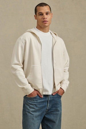 A person stands facing forward, hands in pockets, wearing the VINCENT HOODIE by Velvet by Graham & Spencer over a white tee and blue jeans, set against a neutral background.