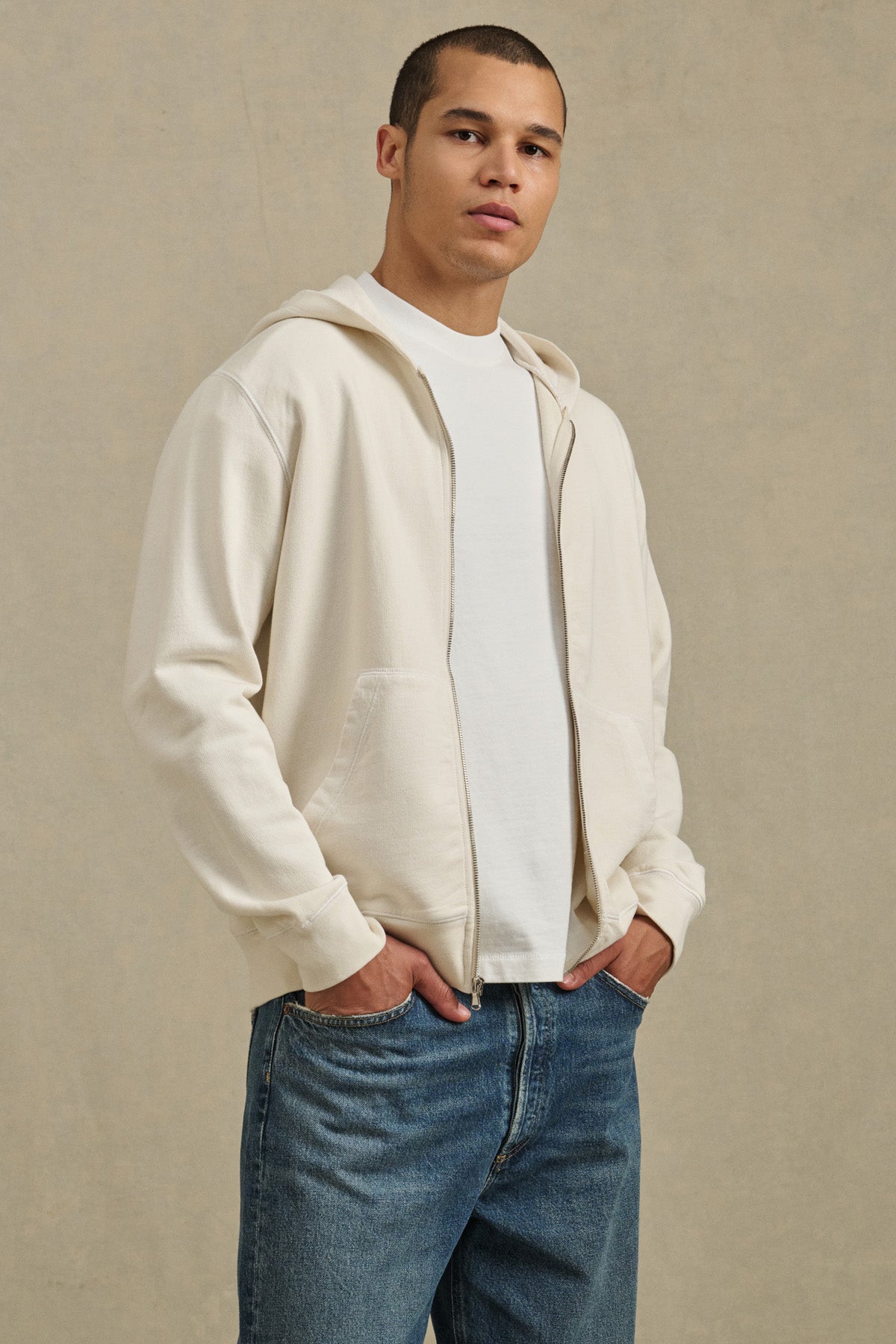 A person models the VINCENT HOODIE by Velvet by Graham & Spencer—a vintage-inspired French terry cotton full-zip style—layered over a white T-shirt and blue jeans, standing with hands in pockets against a neutral background.-41374413914305