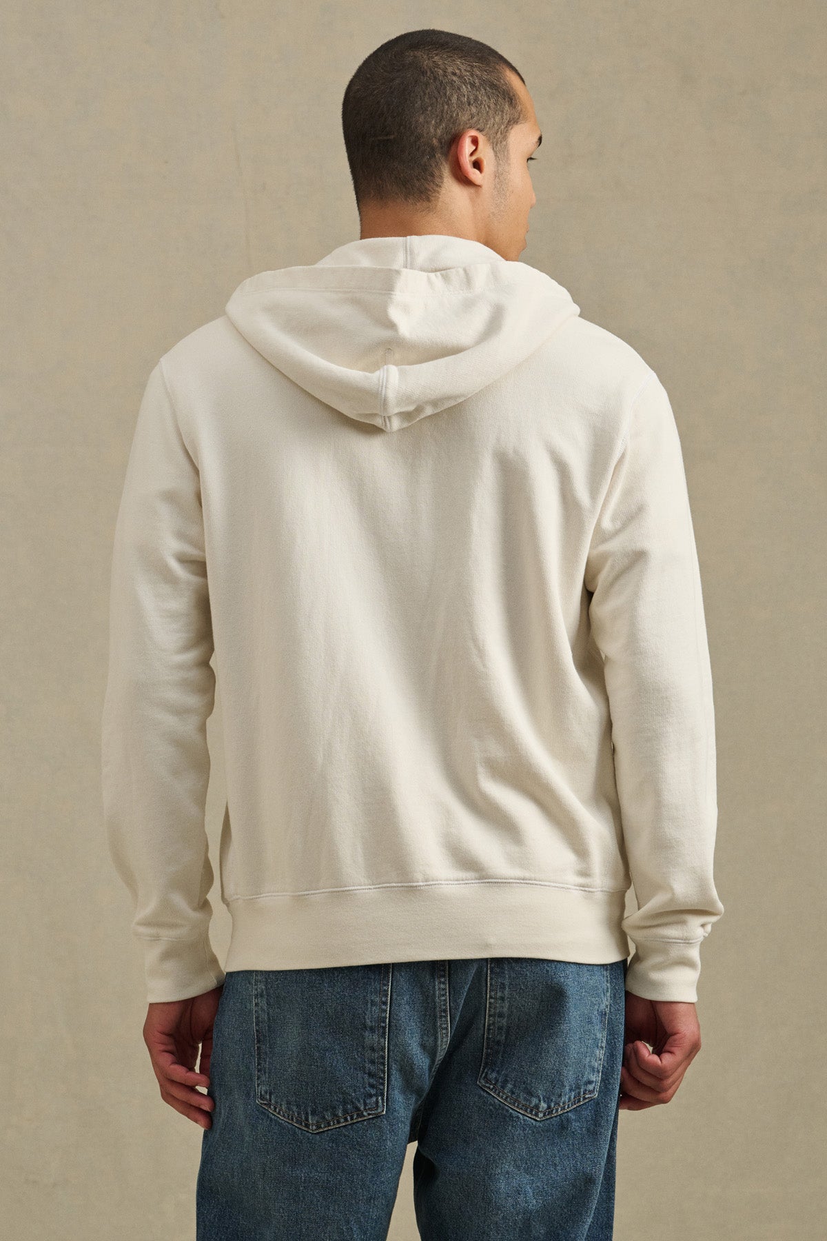 A person in the Velvet by Graham & Spencer VINCENT HOODIE, a vintage-inspired, light-colored French terry cotton full-zip, and blue jeans stands facing away from the camera against a plain backdrop.