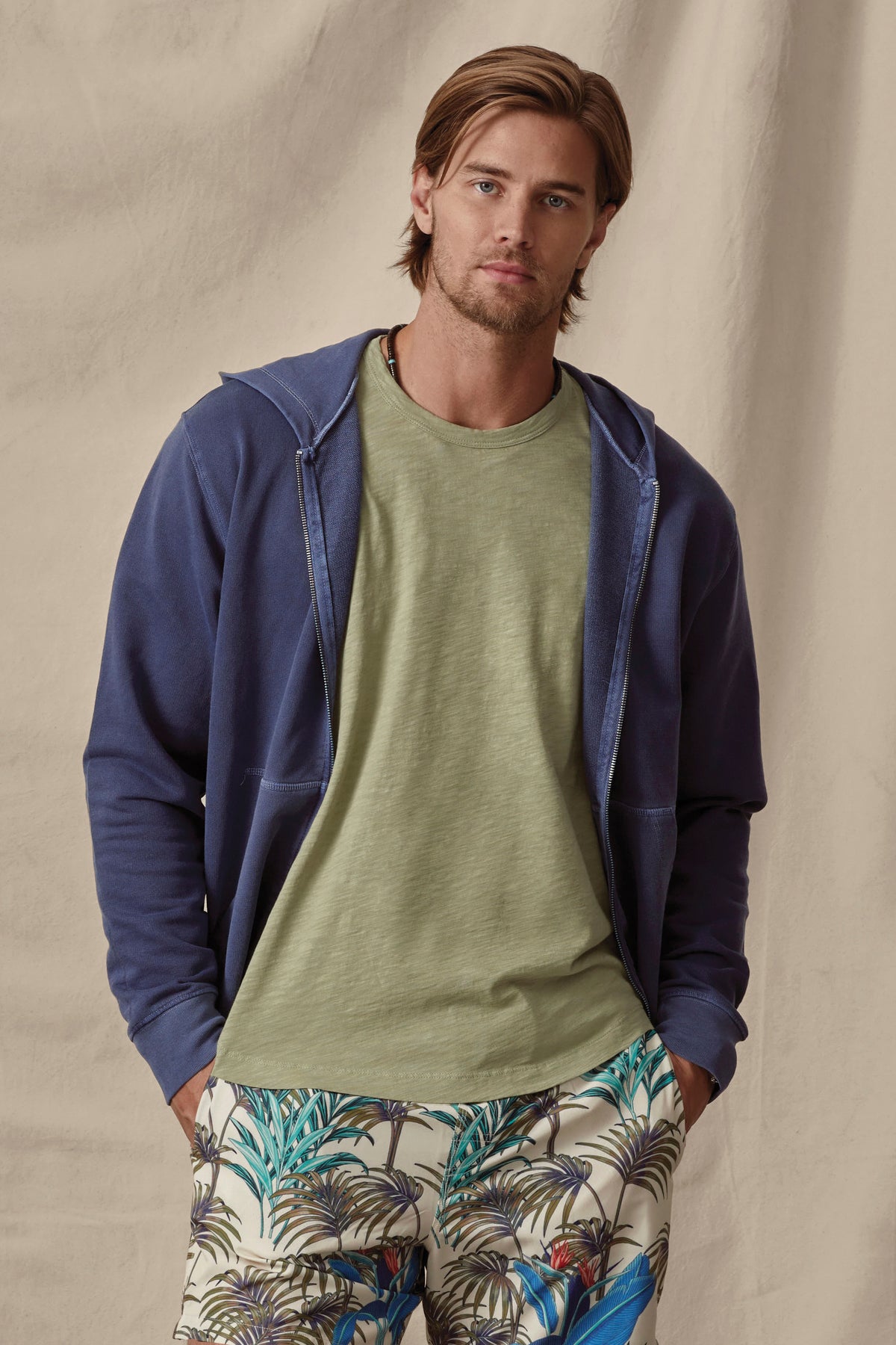 A man with shoulder-length hair wearing a Velvet by Graham & Spencer Vincent Hoodie, green t-shirt, and tropical print pants, standing against a beige backdrop.-36732538290369