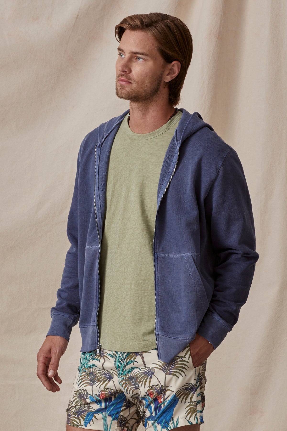 Man in a casual outfit featuring a Velvet by Graham & Spencer VINCENT HOODIE, green t-shirt, and tropical print shorts, standing against a beige backdrop.-36732533113025
