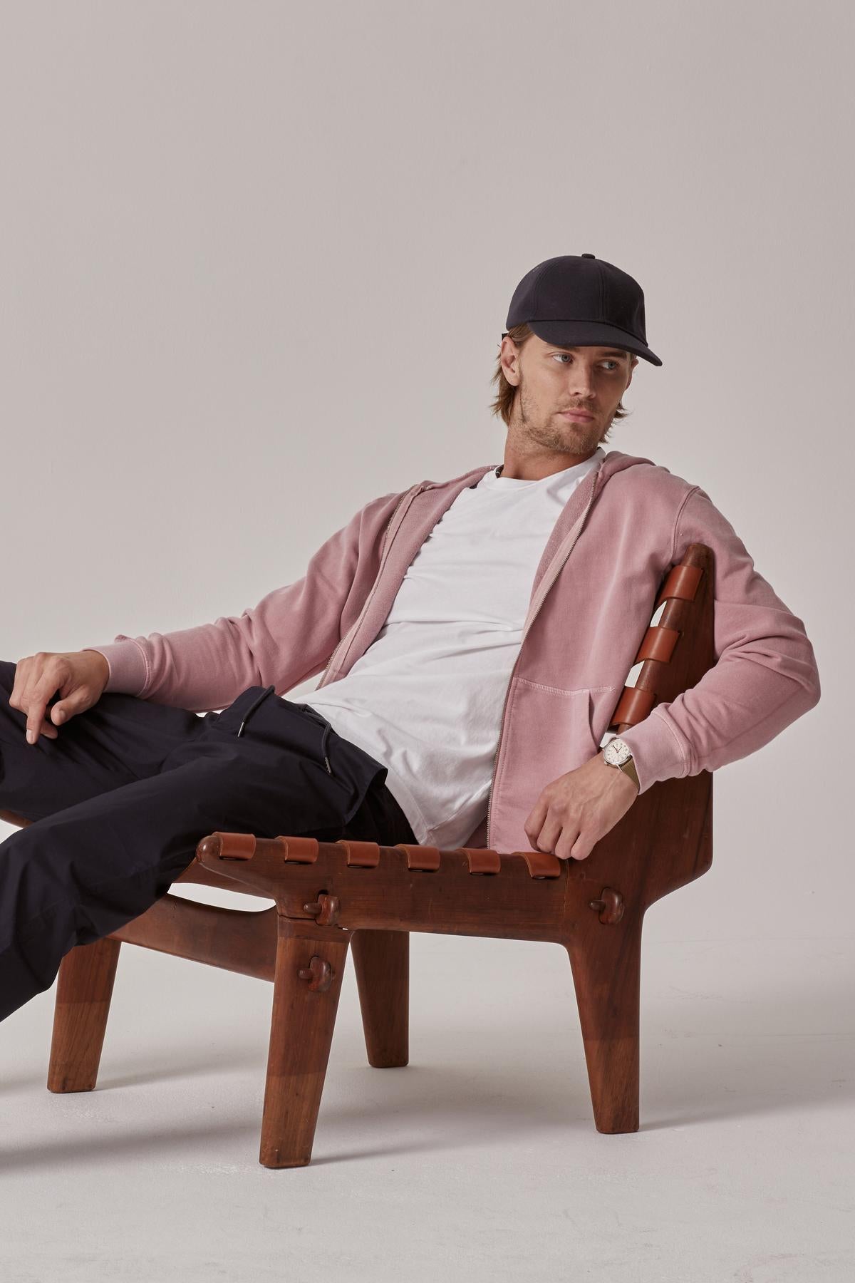   A man in a pink jacket, white t-shirt, and black pants sits casually on a wooden chair, wearing a black cap and looking towards the camera wearing the Velvet by Graham & Spencer VINCENT HOODIE. 