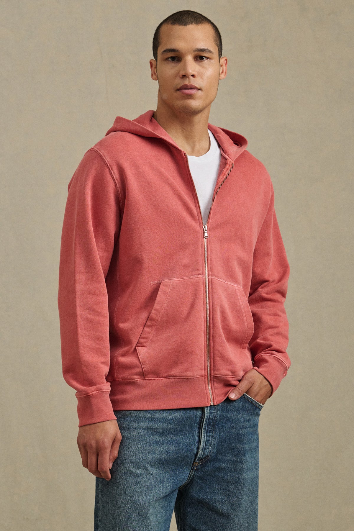 A man stands against a beige background wearing Velvet by Graham & Spencer’s VINCENT HOODIE, a vintage-inspired pink full-zip French terry cotton layer, styled over a white t-shirt and blue jeans.