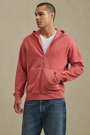 A person stands against a plain background wearing the VINCENT HOODIE by Velvet by Graham & Spencer—a vintage-inspired, faded red full-zip hoodie—over a white t-shirt and blue jeans.