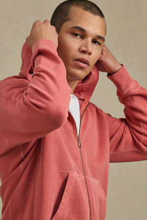 A person wearing the VINCENT HOODIE by Velvet by Graham & Spencer in vintage-inspired faded red, hood up and adjusting it with both hands, stands against a neutral background.