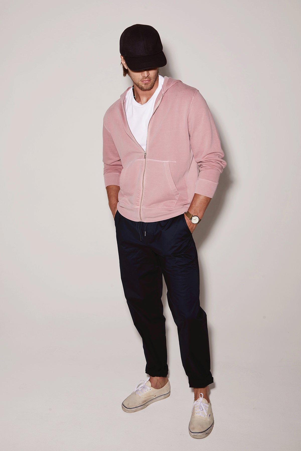 A man in a pink hoodie, white t-shirt, and navy trousers, wearing a black cap and white sneakers, poses against a light background wearing the Velvet by Graham & Spencer LAZARUS JOGGER.-36732528787649