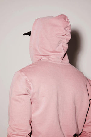 Rear view of a person wearing a Velvet by Graham & Spencer VINCENT HOODIE with the hood up, standing against a light pink wall.
