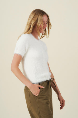 A smiling person wearing a soft, airy white GABBY FEATHER YARN CARDIGAN with short sleeves and olive green pants.