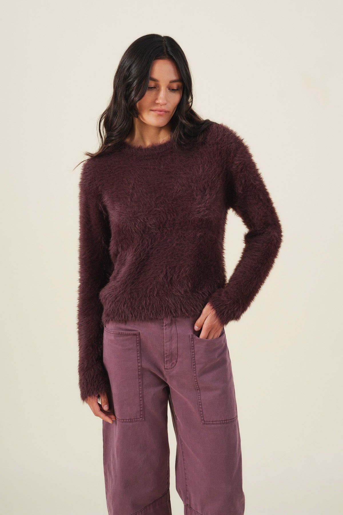 A model wears a long-sleeved, fuzzy dark plum PENNY FEATHER YARN SWEATER by Velvet by Graham & Spencer with a crew neckline and matching plum utility pants for versatile comfort.