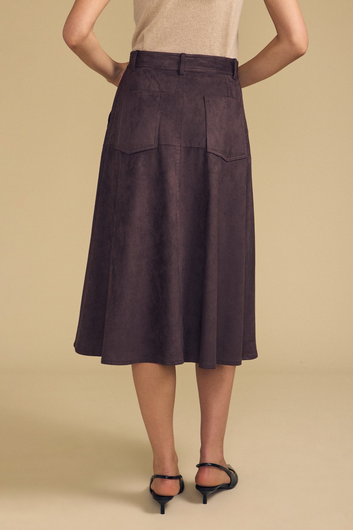 A woman stands with her back to the camera in a beige top and the BELINDA FAUX SUEDE SKIRT by Velvet by Graham & Spencer—its dark brown faux suede and back pockets paired with black heeled sandals create an effortlessly chic transitional look against a neutral backdrop.