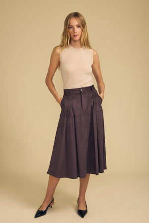 A woman stands against a plain beige background, exuding effortless transitional style in the BELINDA FAUX SUEDE SKIRT by Velvet by Graham & Spencer—a high-waisted, knee-length plum skirt with pleats—paired with a sleeveless beige top and black pointed-toe heels.