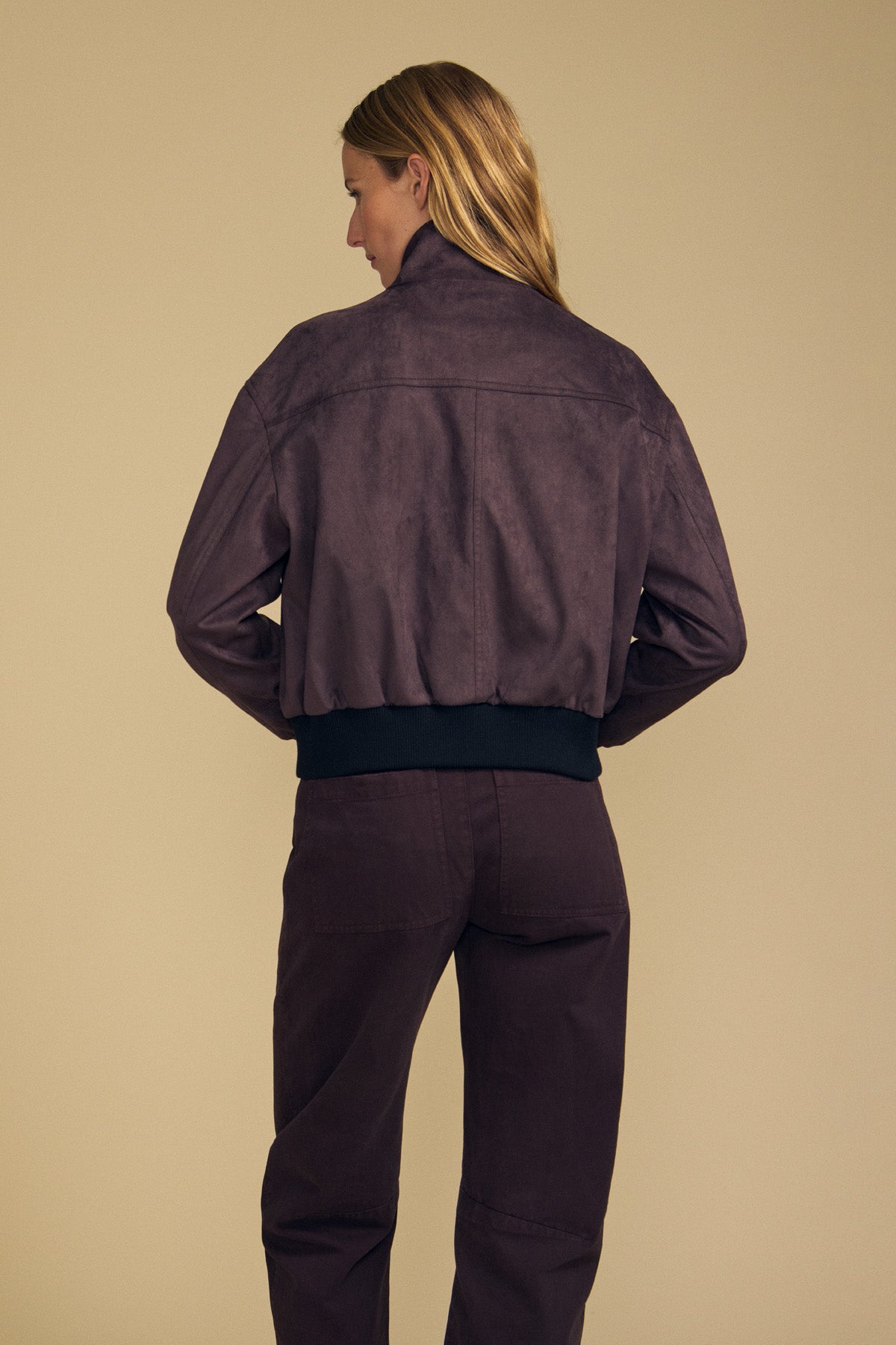 A woman stands facing away, wearing the Velvet by Graham & Spencer QUINCY FAUX SUEDE BOMBER JACKET in purple with a raised collar and matching purple pants, highlighting a modern silhouette against a plain beige background.