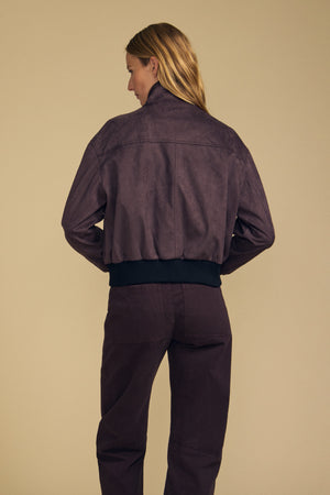 A woman stands facing away, wearing the Velvet by Graham & Spencer QUINCY FAUX SUEDE BOMBER JACKET in purple with a raised collar and matching purple pants, highlighting a modern silhouette against a plain beige background.