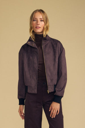 A woman stands against a beige background, wearing the QUINCY FAUX SUEDE BOMBER JACKET by Velvet by Graham & Spencer over a brown turtleneck and matching brown pants, creating a chic modern silhouette.