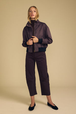 A woman wears the QUINCY FAUX SUEDE BOMBER JACKET by Velvet by Graham & Spencer in brown, paired with matching wide-leg pants, a black turtleneck, and black flats, standing against a plain beige background.